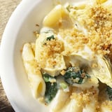 A close-up of a serving of Artichoke Pasta Bake reveals tender pasta, wilted spinach, and marinated artichoke hearts in a rich, cheesy cream sauce.  