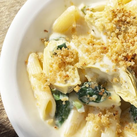 A close-up of a serving of Artichoke Pasta Bake reveals tender pasta, wilted spinach, and marinated artichoke hearts in a rich, cheesy cream sauce.  