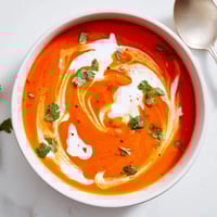 A bowl of Roasted Red Pepper Soup with swirls of crème fraîche and fresh parsley garnish, served next to rustic crusty bread.